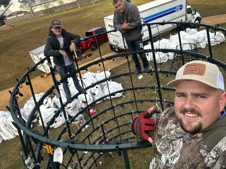 Three crew members assembling the steel ring frame of a commercial Christmas tree on a lawn in daylight, one taking a photograph in the foreground.