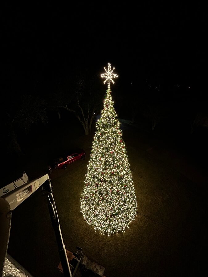 A tall commercial display tree lit in warm white with a lit star topper, seen at night from the basket of a lift.