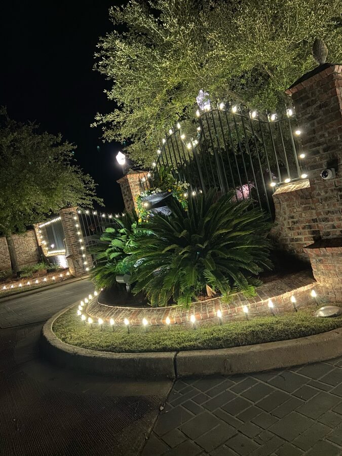 An iron entrance gate between brick pillars at night, its arch outlined in warm white bulbs and the landscape bed below edged with lights.
