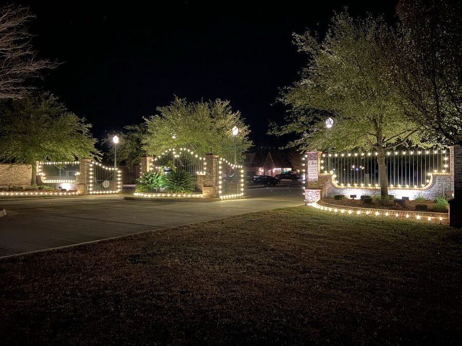 Perimeter walls and iron gates outlined in warm white lights at a property entrance at night.