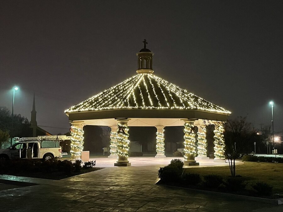 Wide night view of a lit public pavilion with wrapped columns and an outlined roof.