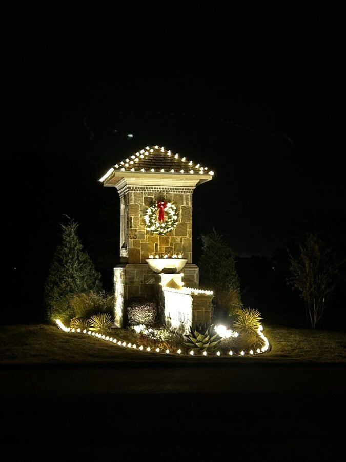 A lit stone entrance column with a wreath and surrounding landscape lighting after dark.