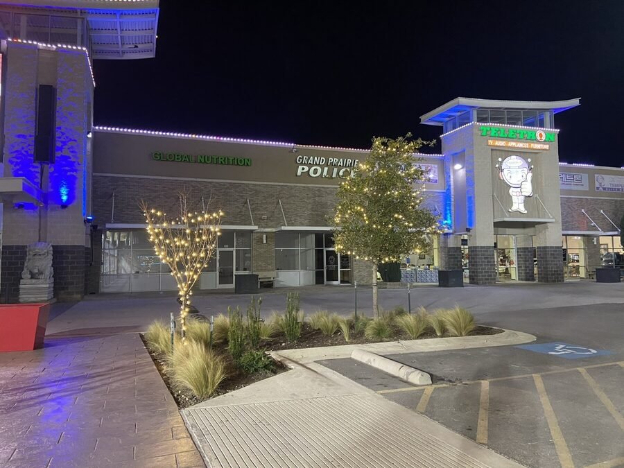 Wide night view of a retail center facade with wrapped landscape trees along the storefronts.